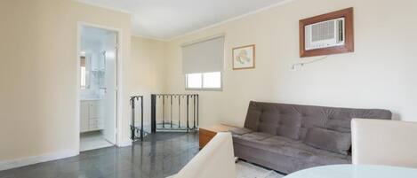 Una sala moderna con un sofá gris, una mesa de centro de vidrio y una mesa de comedor blanca. Hay una pequeña mesita de madera y un soporte de metal negro. La habitación tiene un ventanal grande con una persiana blanca, paredes beige y una lámpara de techo.