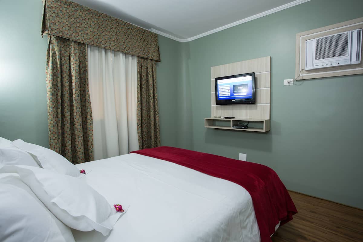 A hotel room with a bed, curtains, a television mounted on the wall, and a small shelf below it.