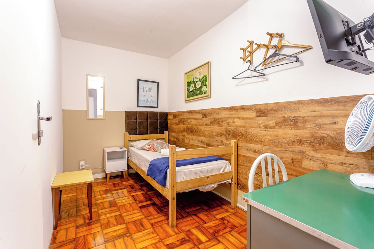 A small, neatly arranged room with a wooden bed, a small desk, a chair, and a wall-mounted fan.