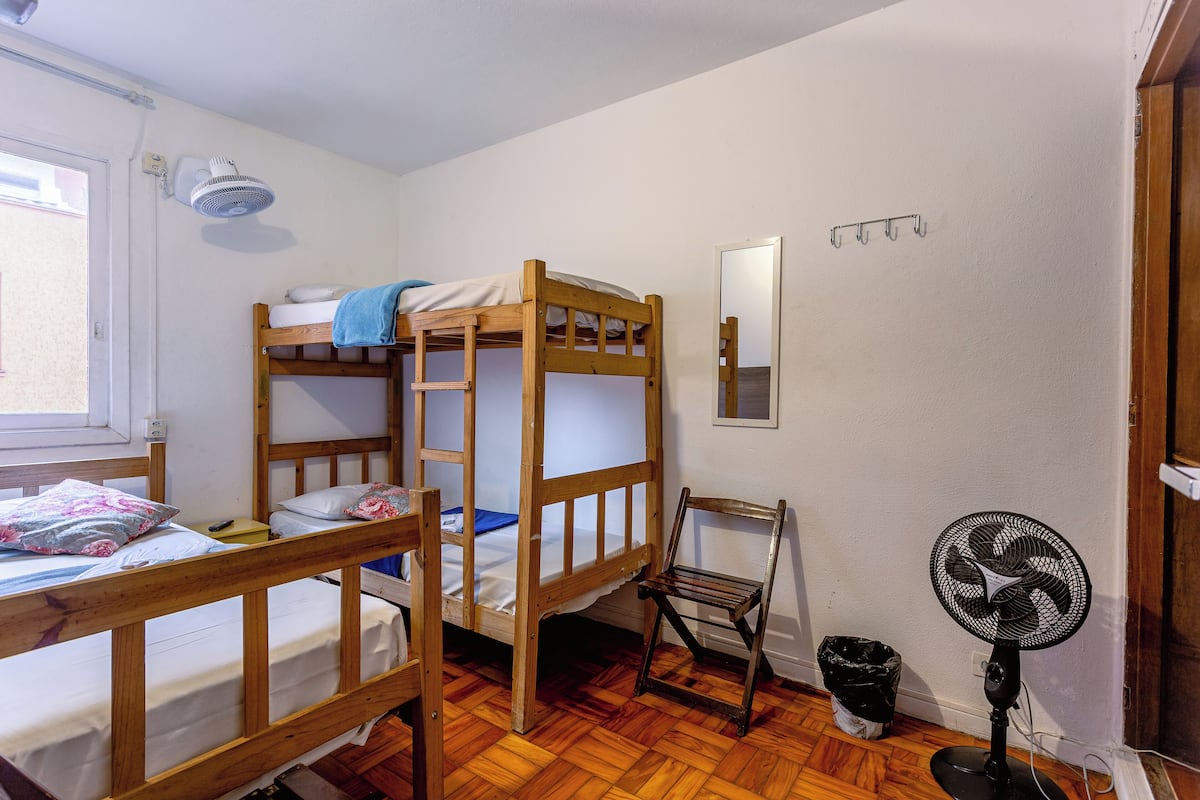 A bunk bed room with a wooden floor, a desk, a chair, a standing fan, and a window.