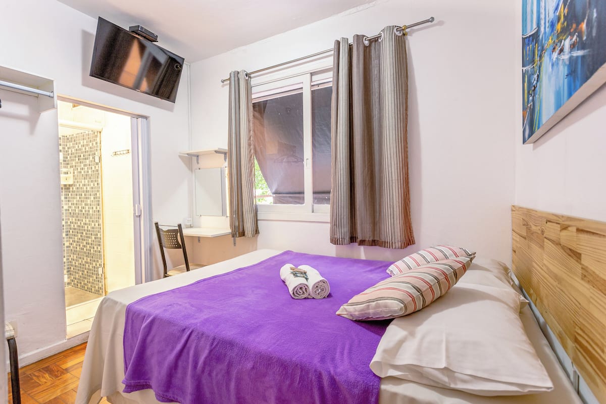A bedroom with a bed, a TV mounted on the wall, a window with curtains, and a painting on the wall.