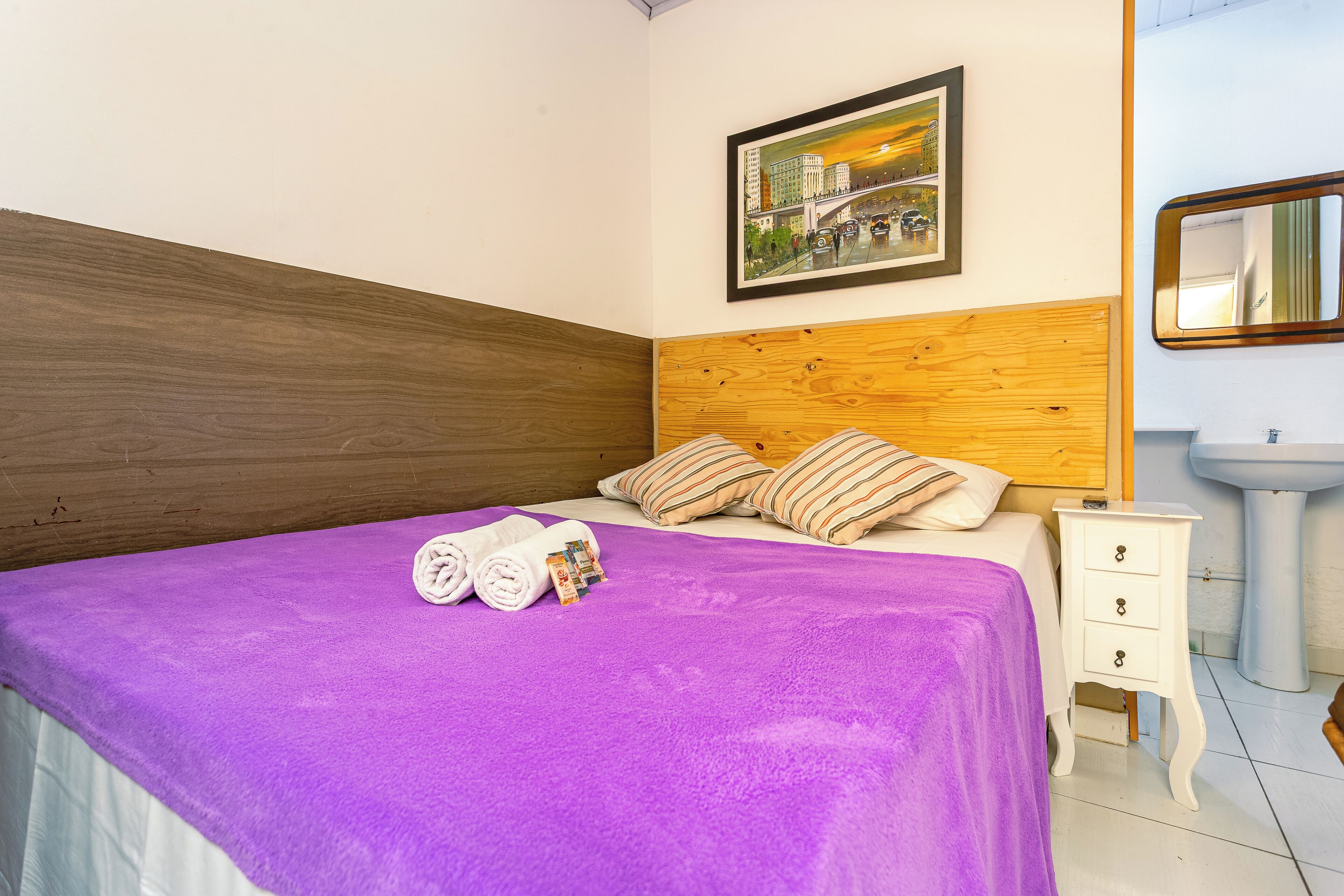 Una cama bien tendida con una colcha púrpura, un cabecero de madera y almohadas a rayas. Hay una mesita de noche pequeña con un cajón y un espejo montado en la pared. Una foto enmarcada cuelga sobre la cama.