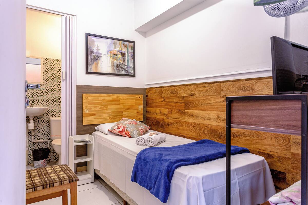 A small, neatly arranged bedroom with a wooden headboard, a bed with a blue blanket, a small bedside table, a chair, and a TV.
