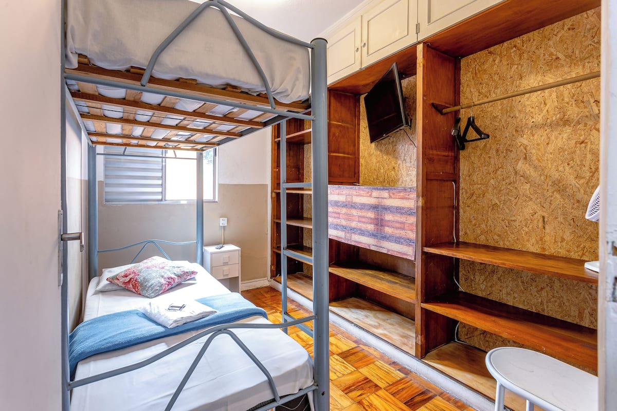 A bunk bed with a wooden headboard, a small bedside table, and a window with blinds.