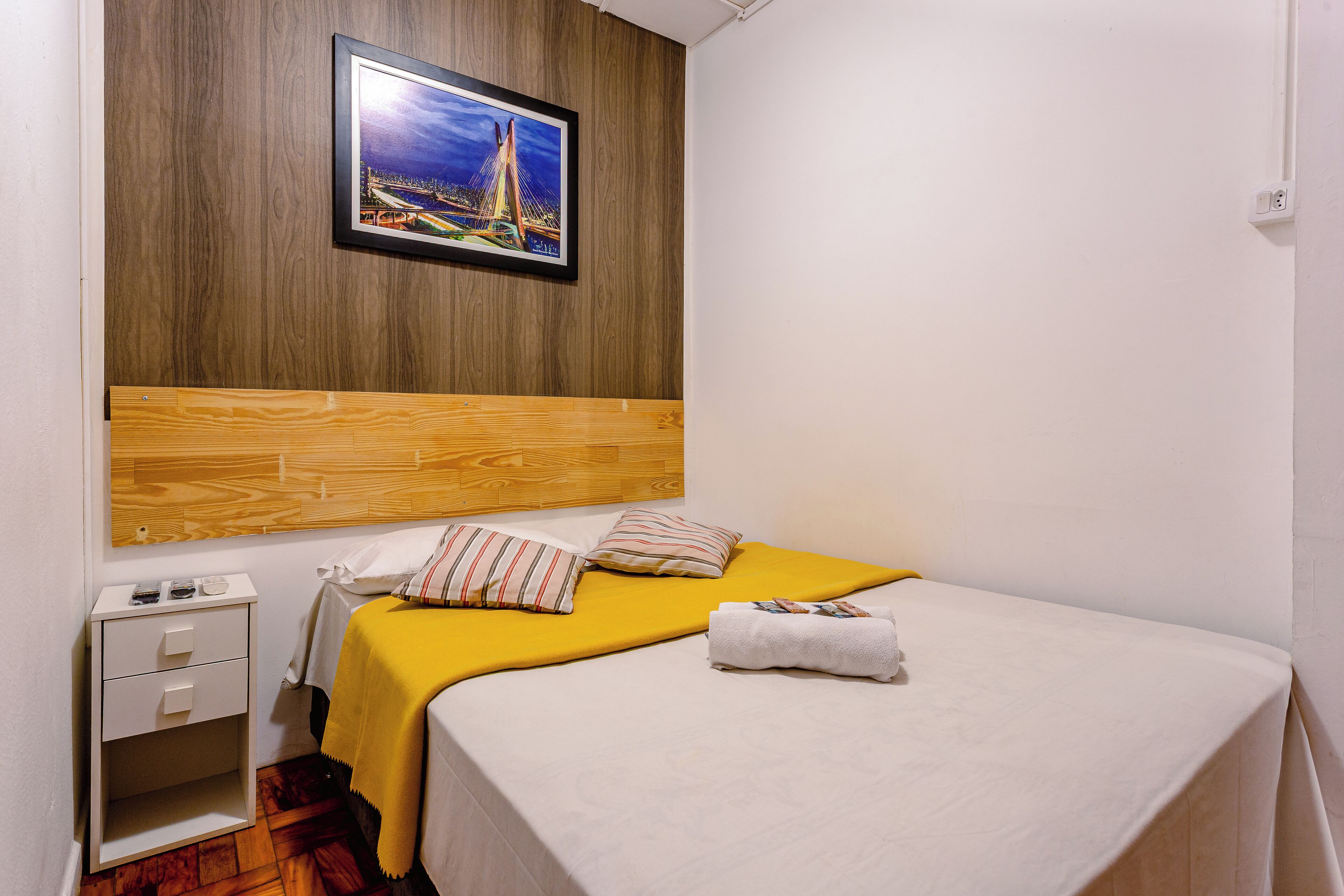 A neatly arranged hotel room with a wooden headboard, a bed with a yellow bedspread, two pillows, a white bedside table with drawers, and a framed picture on the wall.
