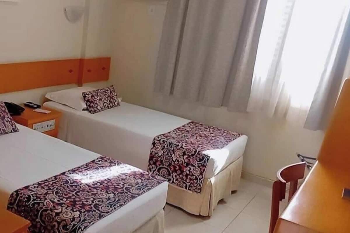 Habitación de hotel con dos camas individuales, una mesita de noche, una silla y un ventanal con cortinas.