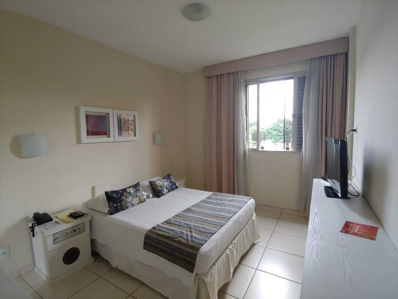 A hotel room with a bed, a bedside table with a phone and a lamp, a TV on a stand, and a window with curtains.