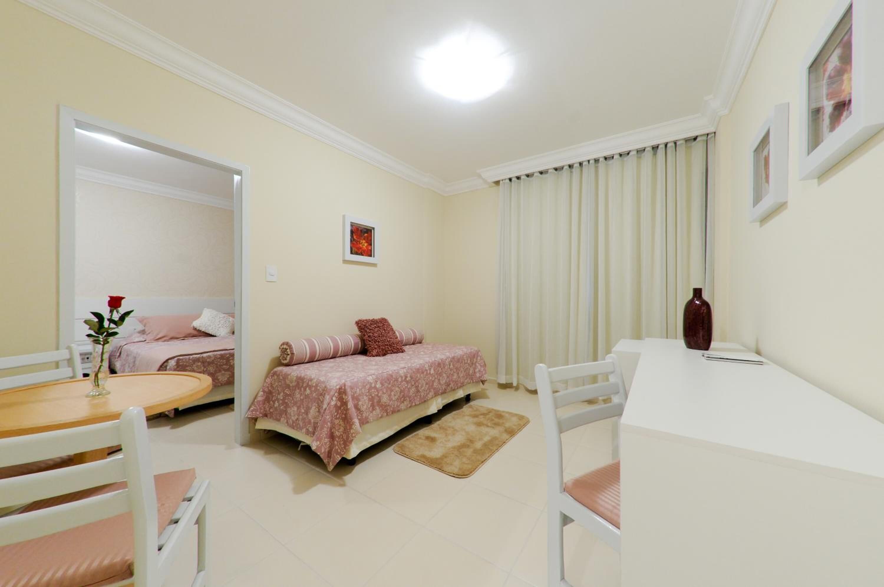 Un dormitorio con cama, mesita de noche, una silla, una pequeña mesa y una ventana con cortinas.