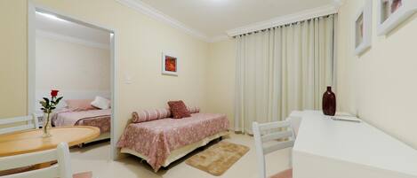 Un dormitorio con cama, mesita de noche, una silla, una pequeña mesa y una ventana con cortinas.