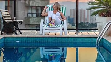 Uma pessoa relaxando em uma cadeira à beira da piscina, com vista para a paisagem urbana ao pôr do sol.