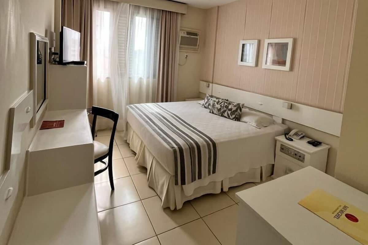 Habitación de hotel con cama, escritorio, silla, mesita de noche con teléfono y ventana con cortinas.