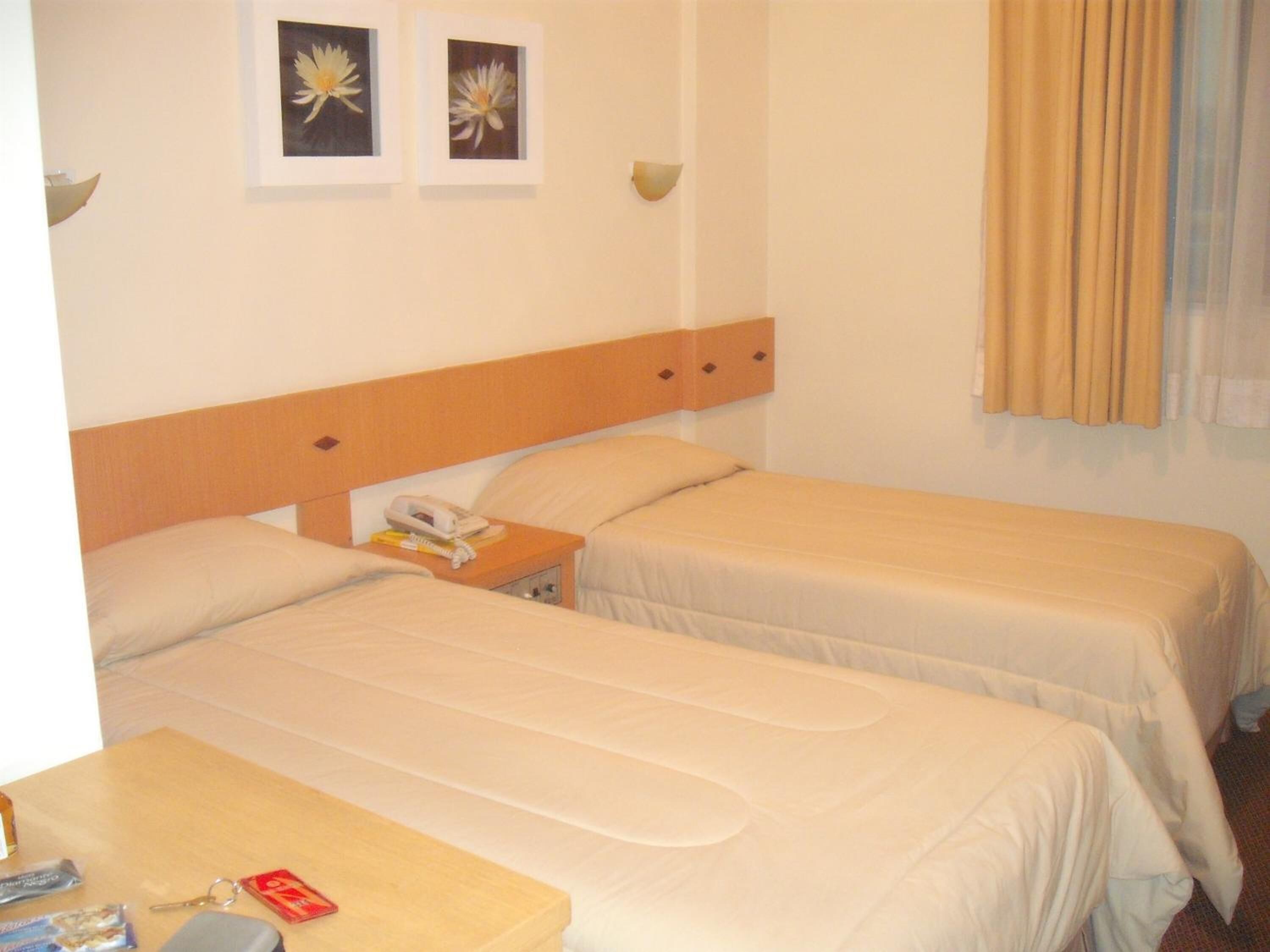 A hotel room with two single beds, a wooden headboard, a bedside table with a phone, and two framed pictures on the wall.