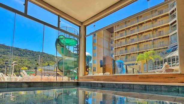 A modern indoor swimming pool with clear glass walls reflecting the surrounding area, including a water slide and a multi-story building with balconies. - Hotel Zanon (Aguas de Lindoia)