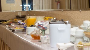 Un bufé con diversos alimentos y bebidas, que incluye un tazón de frutas, un dispensador de jugo y una cafetera.