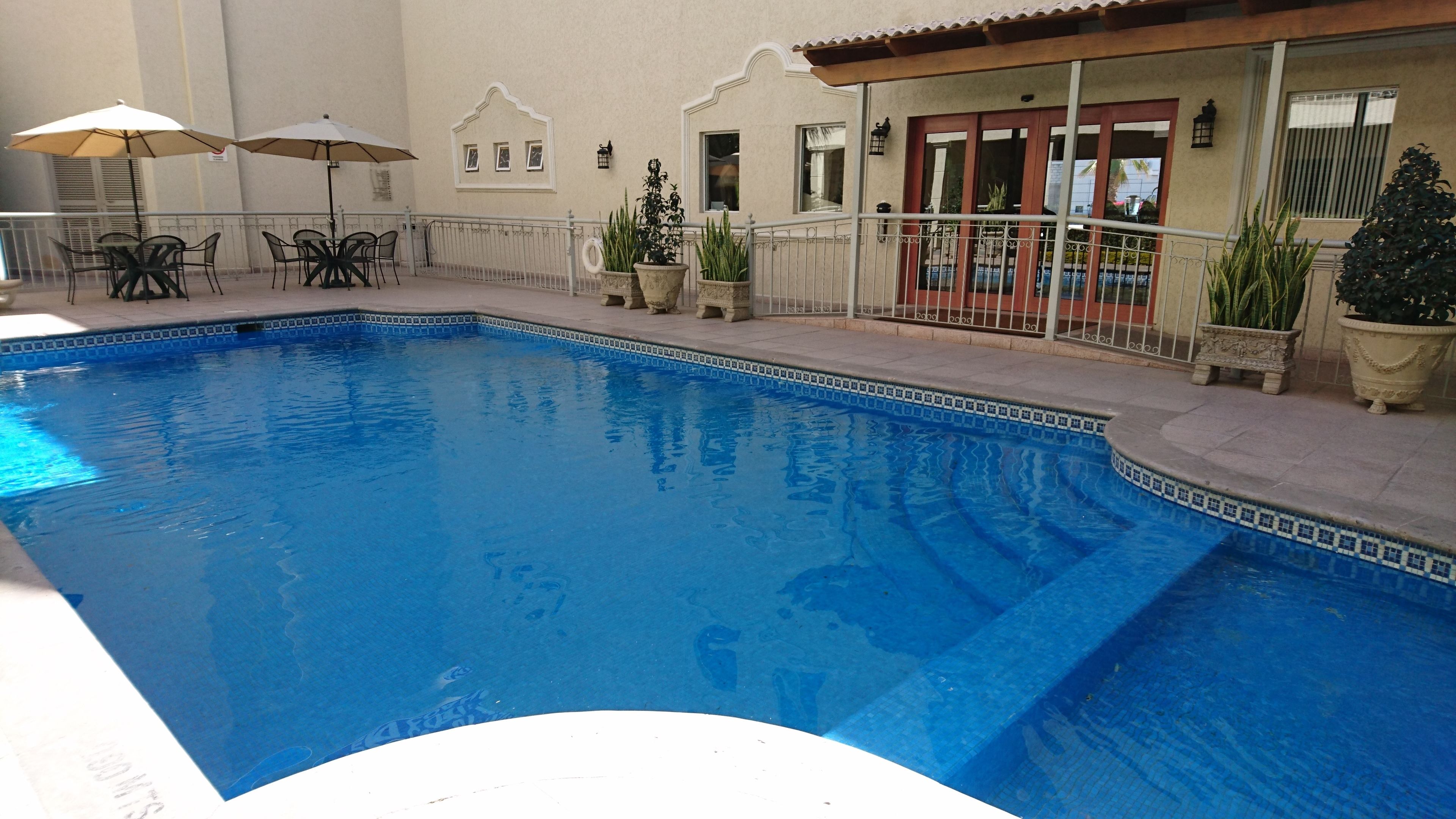 A swimming pool with a tiled edge, surrounded by a patio area with outdoor furniture and potted plants.