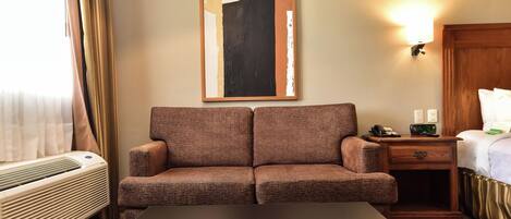 A hotel room with a sofa, a table, a lamp, a painting, and a bed.