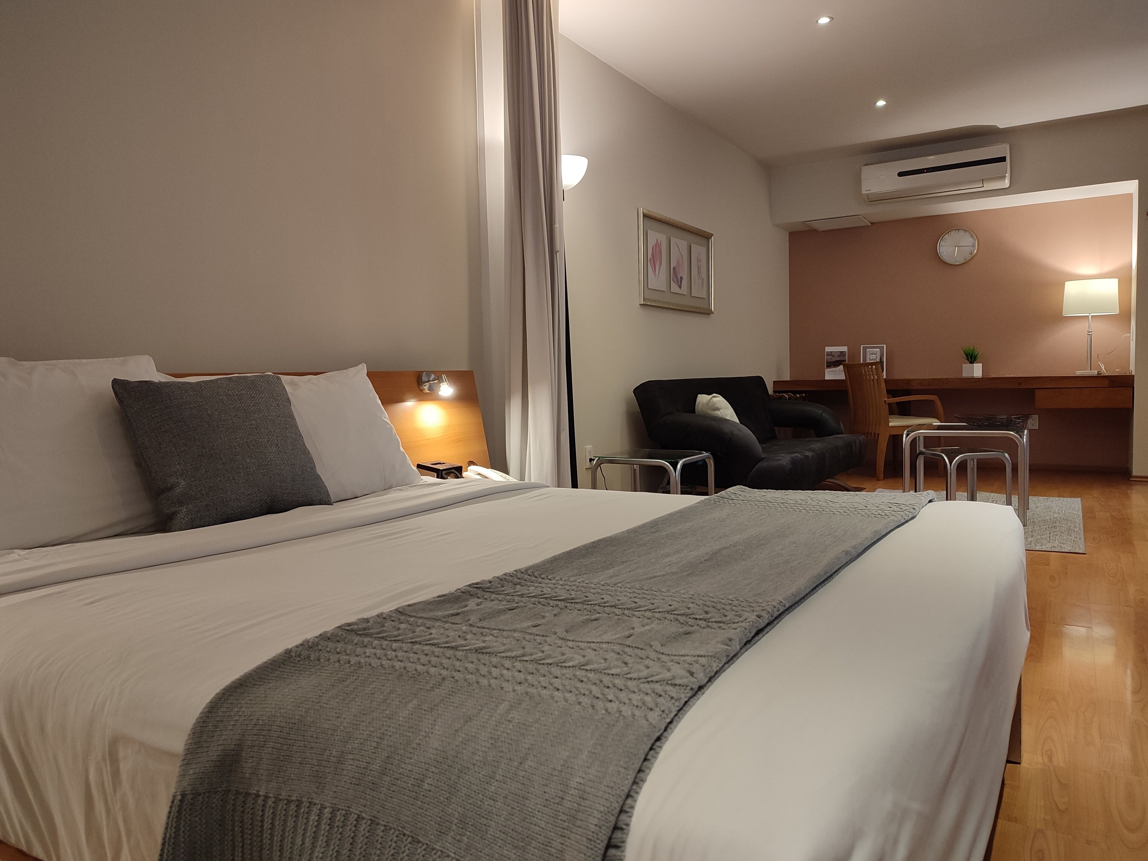 A hotel room with a large bed, a desk, a chair, and an air conditioning unit.