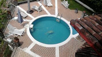 Una piscina circular con borde blanco, rodeada por un patio de ladrillo. Hay sillas reclinables blancas y una pequeña mesa con toldo.