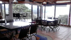 Un área de comedor al aire libre y techada, con una mesa grande y sillas, ventanas amplias y vistas del paisaje.