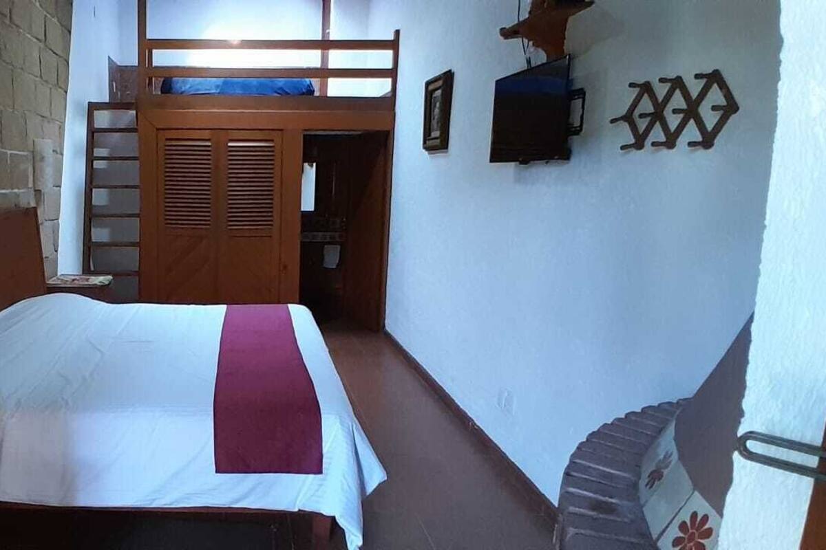 Habitación con una cama individual y cabecera de madera, una cama litera al fondo, una mesita pequeña y un estante fijado a la pared.