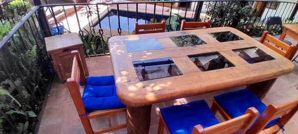 Mesa de madera con paneles de vidrio y cojines azules, rodeada de sillas, en un entorno exterior con piscina y vegetación.