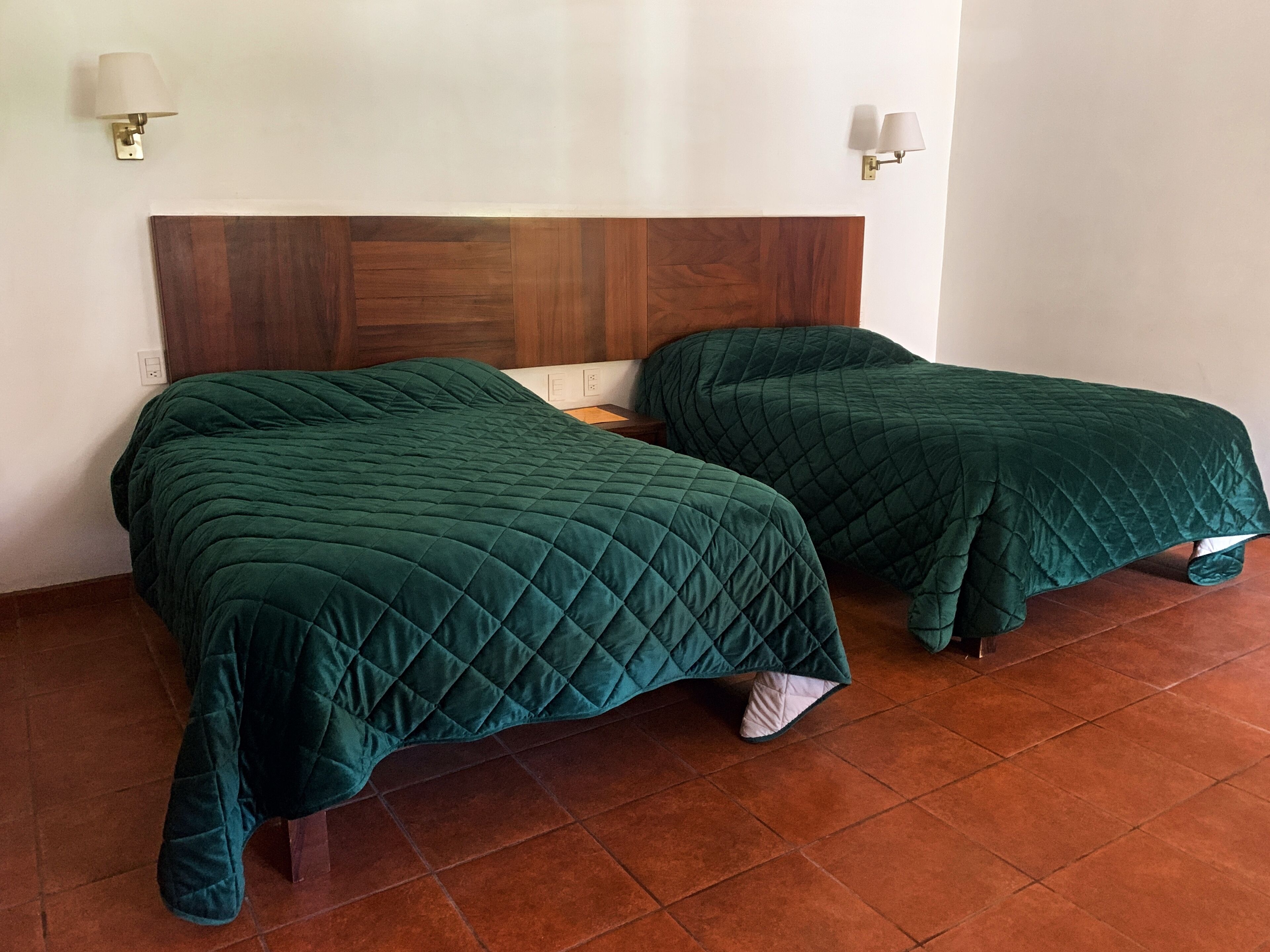 Dos camas con colchas verdes, un cabecero de madera y dos lámparas fijadas a la pared.