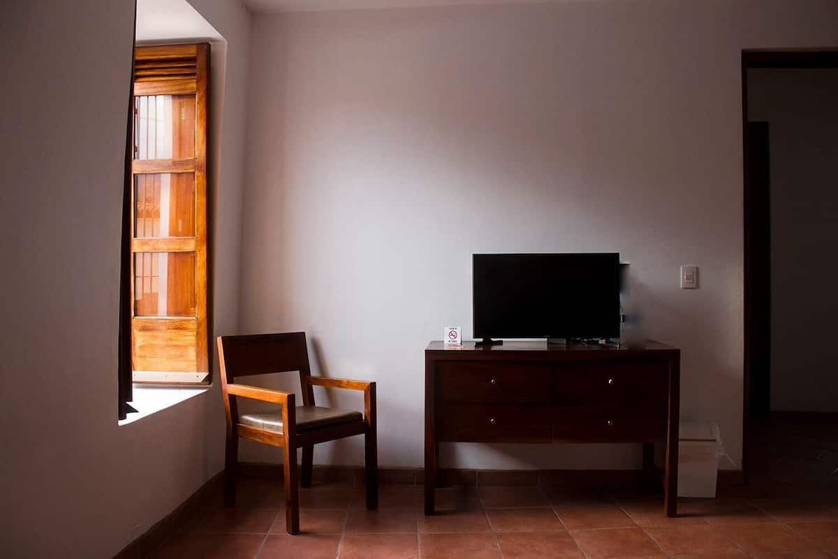 Una habitación con una silla de madera, un televisor sobre un mueble de madera y una ventana con persianas.