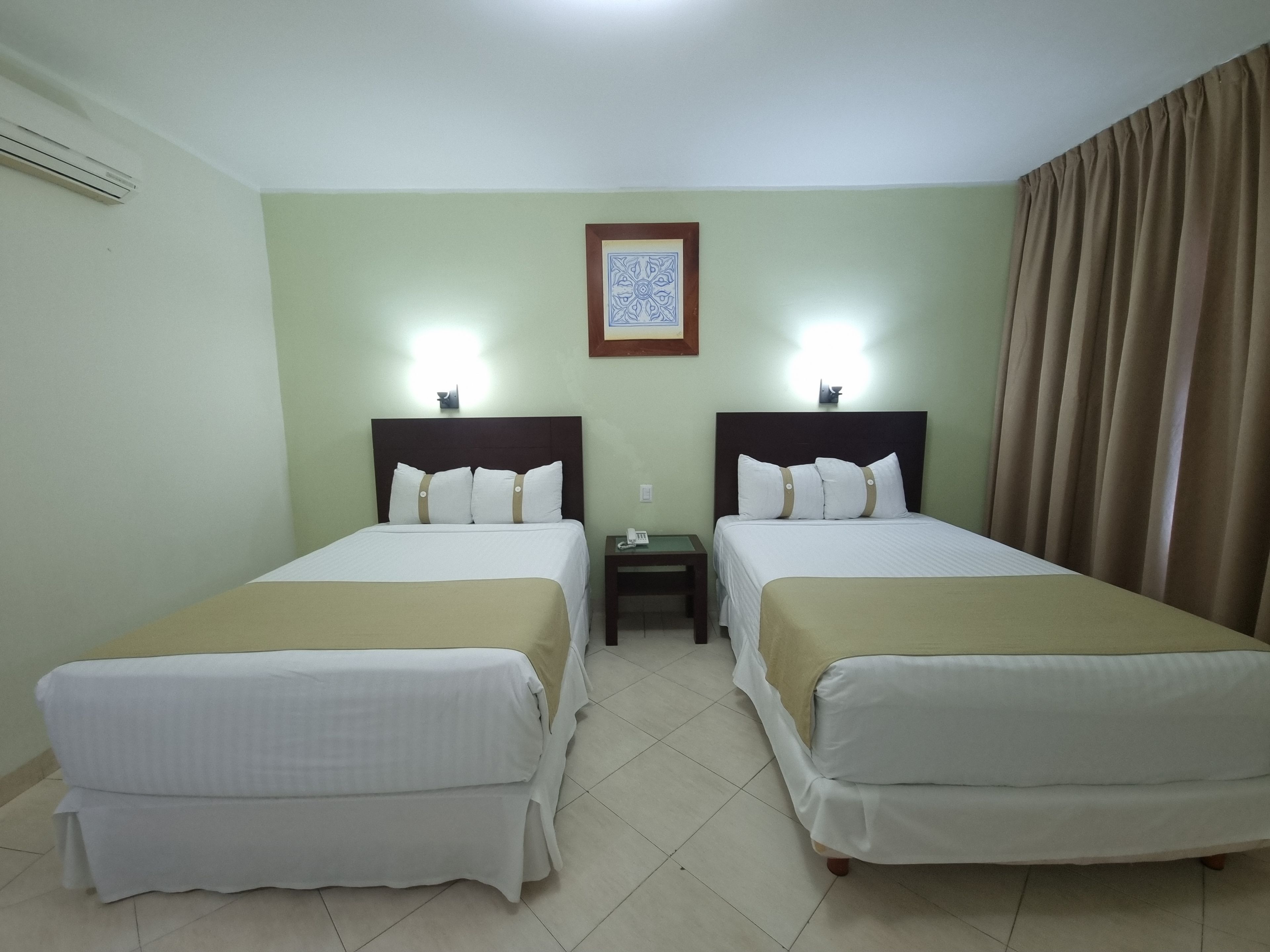A hotel room with two beds, a nightstand, a framed picture on the wall, and a curtain on the window.