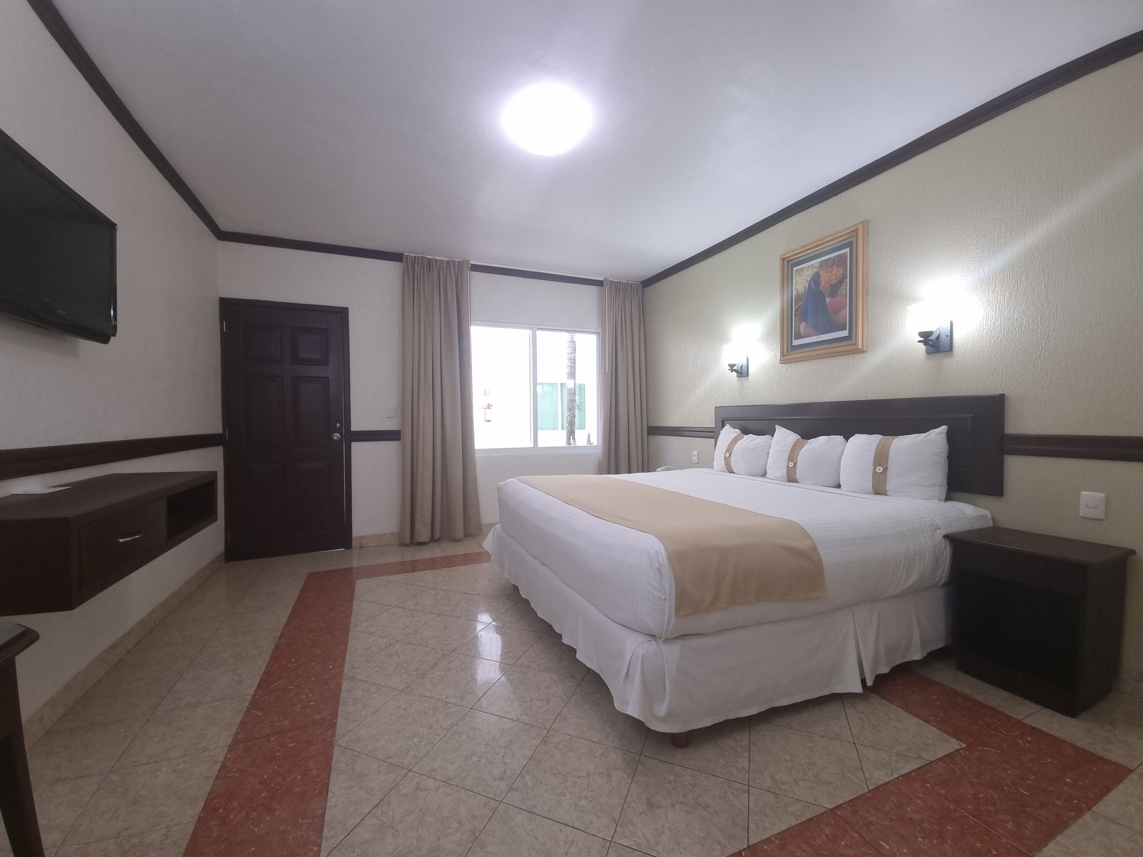A hotel room with a bed, a nightstand, a TV, a window with curtains, and a painting on the wall.