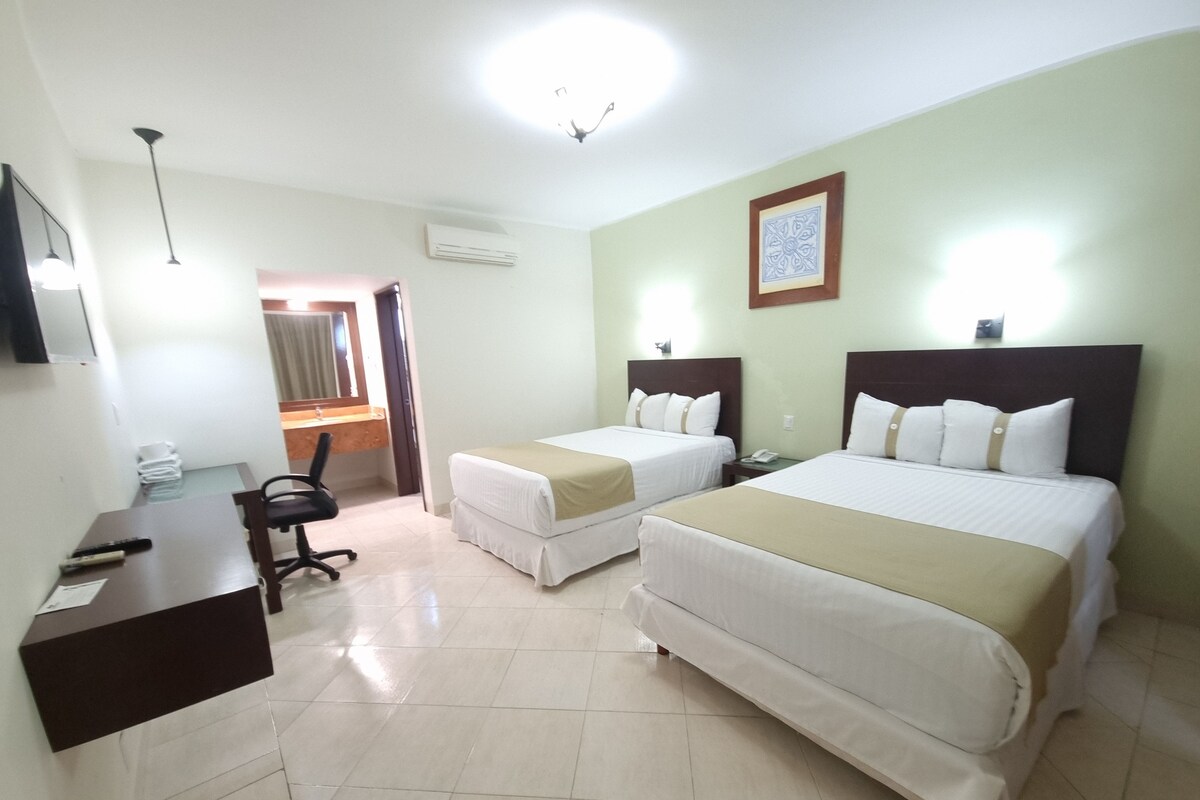 A hotel room with two beds, a desk, a chair, and a bathroom visible through an open door.