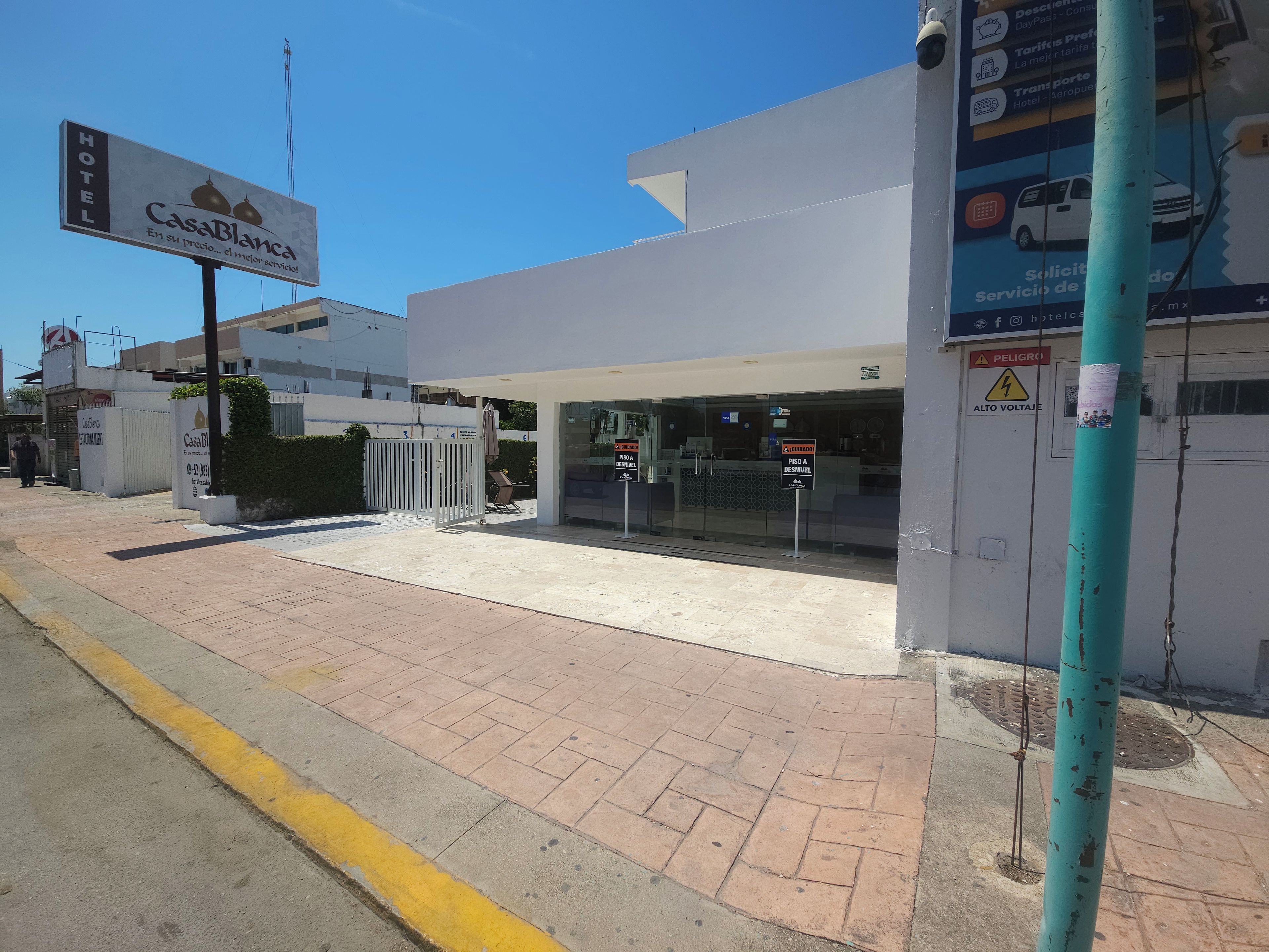Entrada de hotel con un letrero que dice Casa Blanca, acceso mediante portón y señal de advertencia de alta tensión.