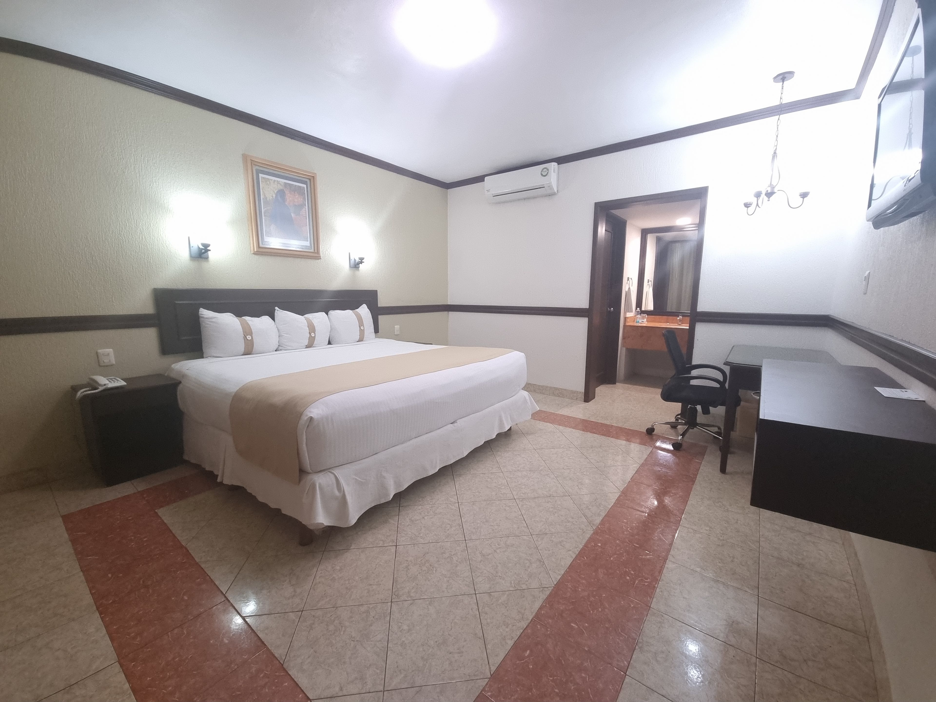 Habitación de hotel con cama, mesita de noche, escritorio, silla y baño visible a través de una puerta abierta.