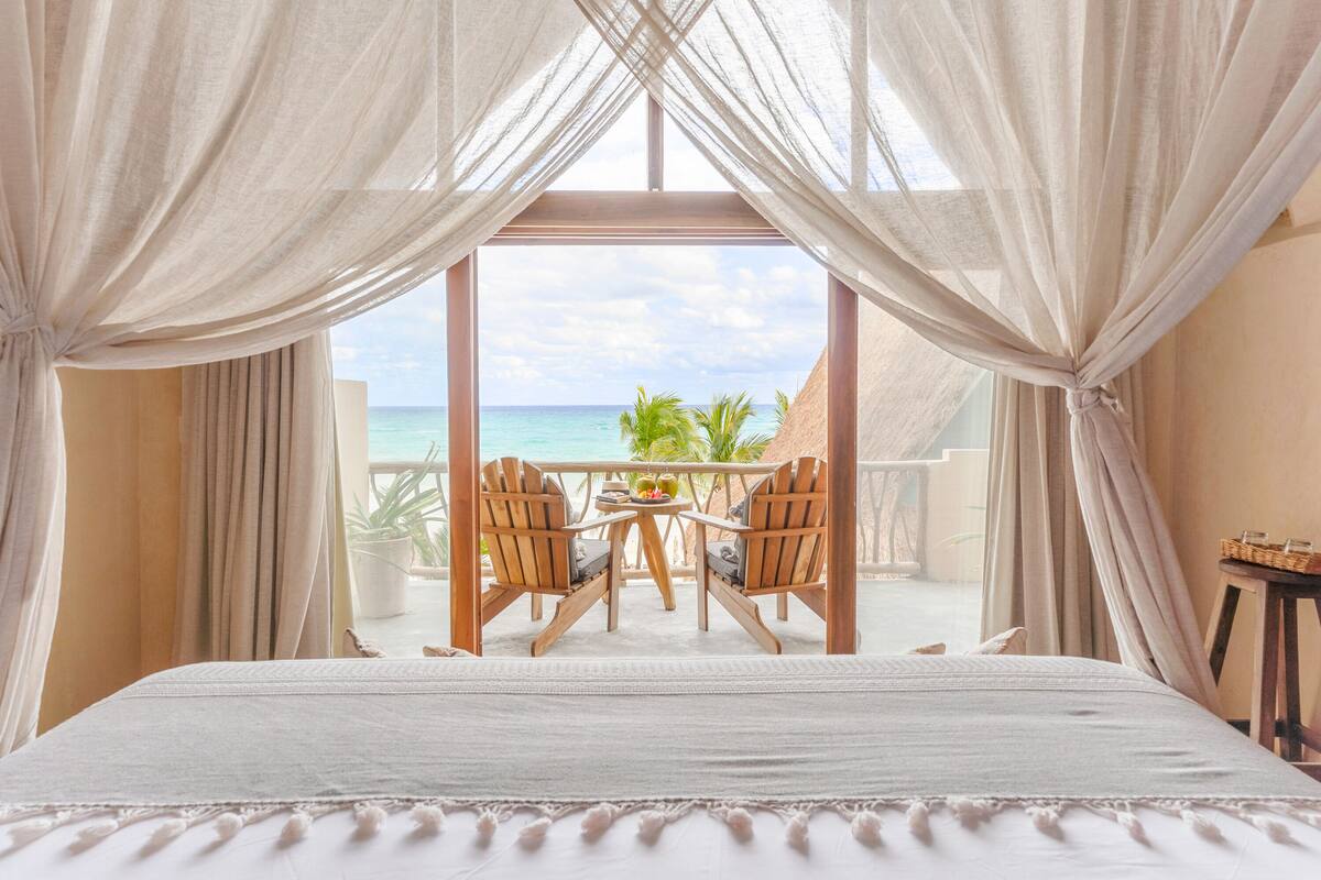A bedroom with a large bed, a balcony with wooden chairs, and a view of the ocean.