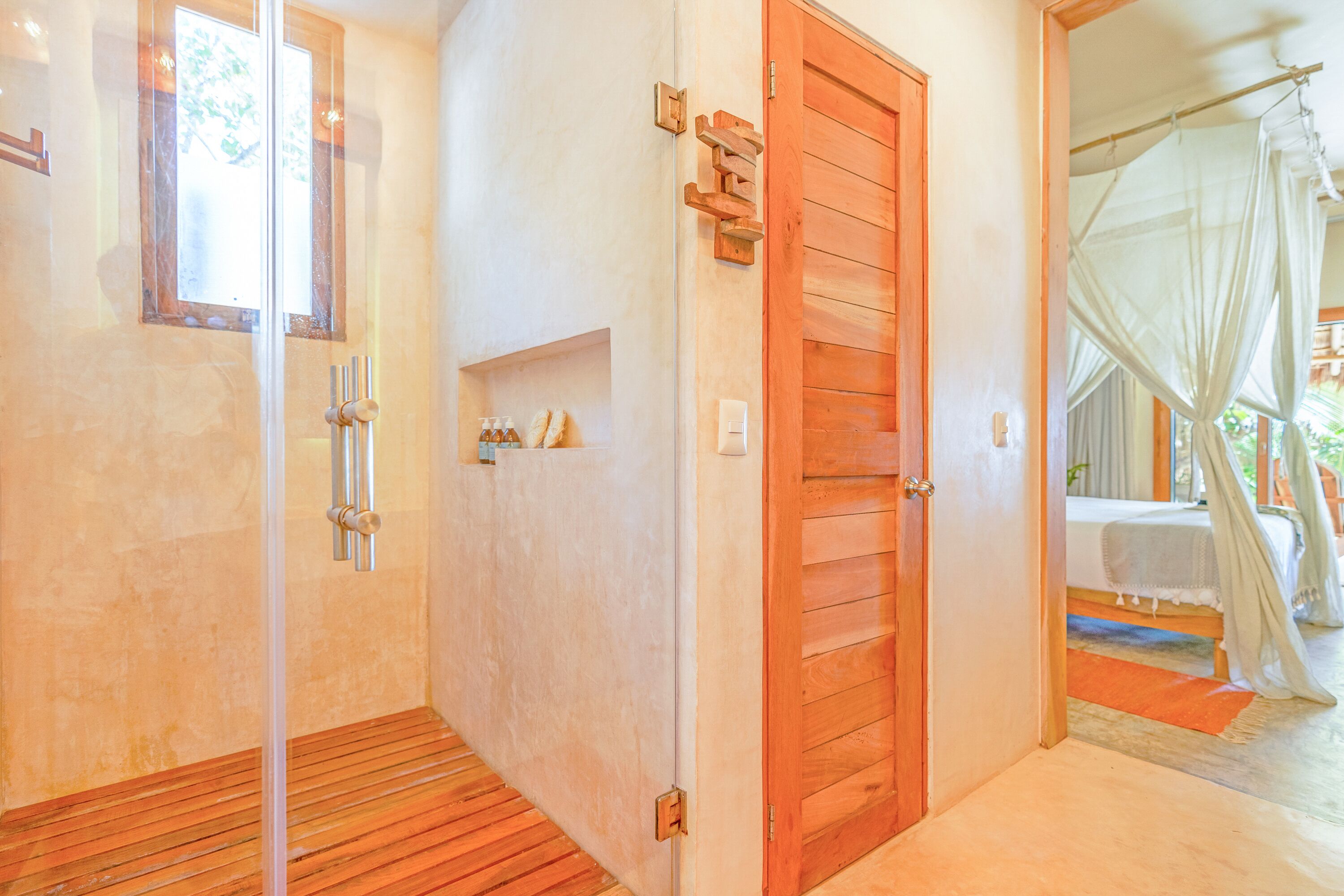 A room with a wooden floor, a wooden door, a shower area, and a bed with a canopy.