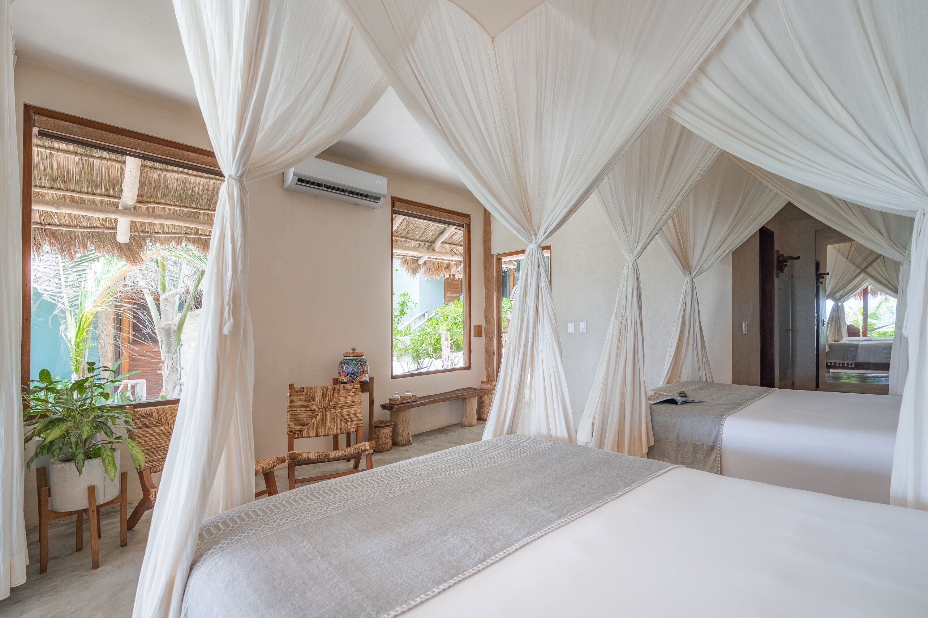 A bedroom with a large bed, a bench, and a chair. There are curtains and a window with a view of palm trees.