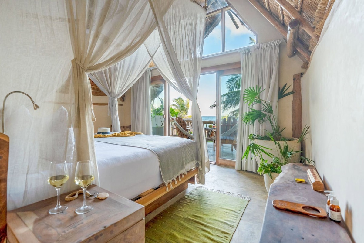 A bedroom with a bed, a nightstand, a table with a bottle and a container, and a view of the ocean.