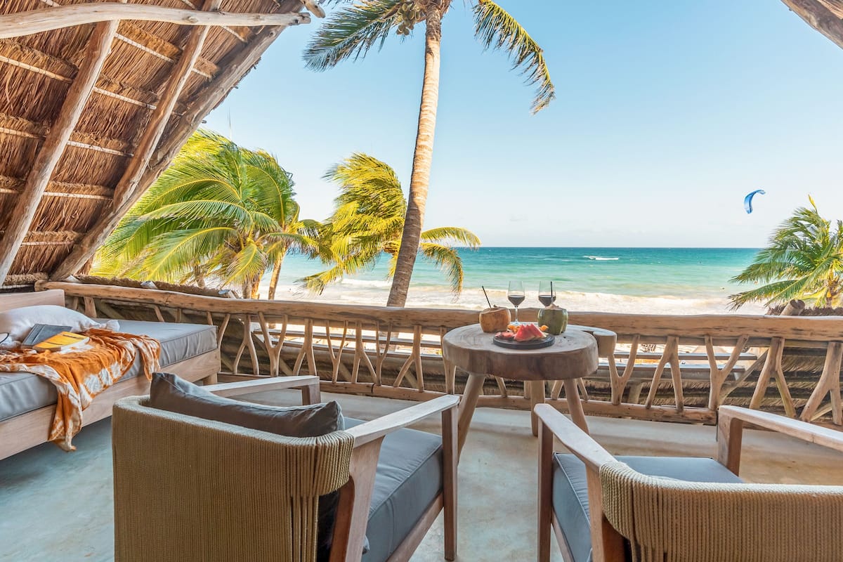 A beachside lounge with a thatched roof, wicker furniture, and a table set with a drink and snacks.