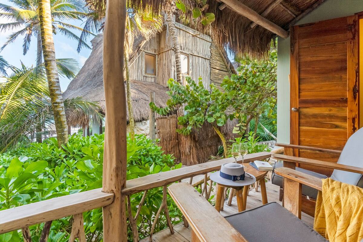 A wooden deck with a thatched-roof structure, a wooden door, and a table with a hat and a glass.