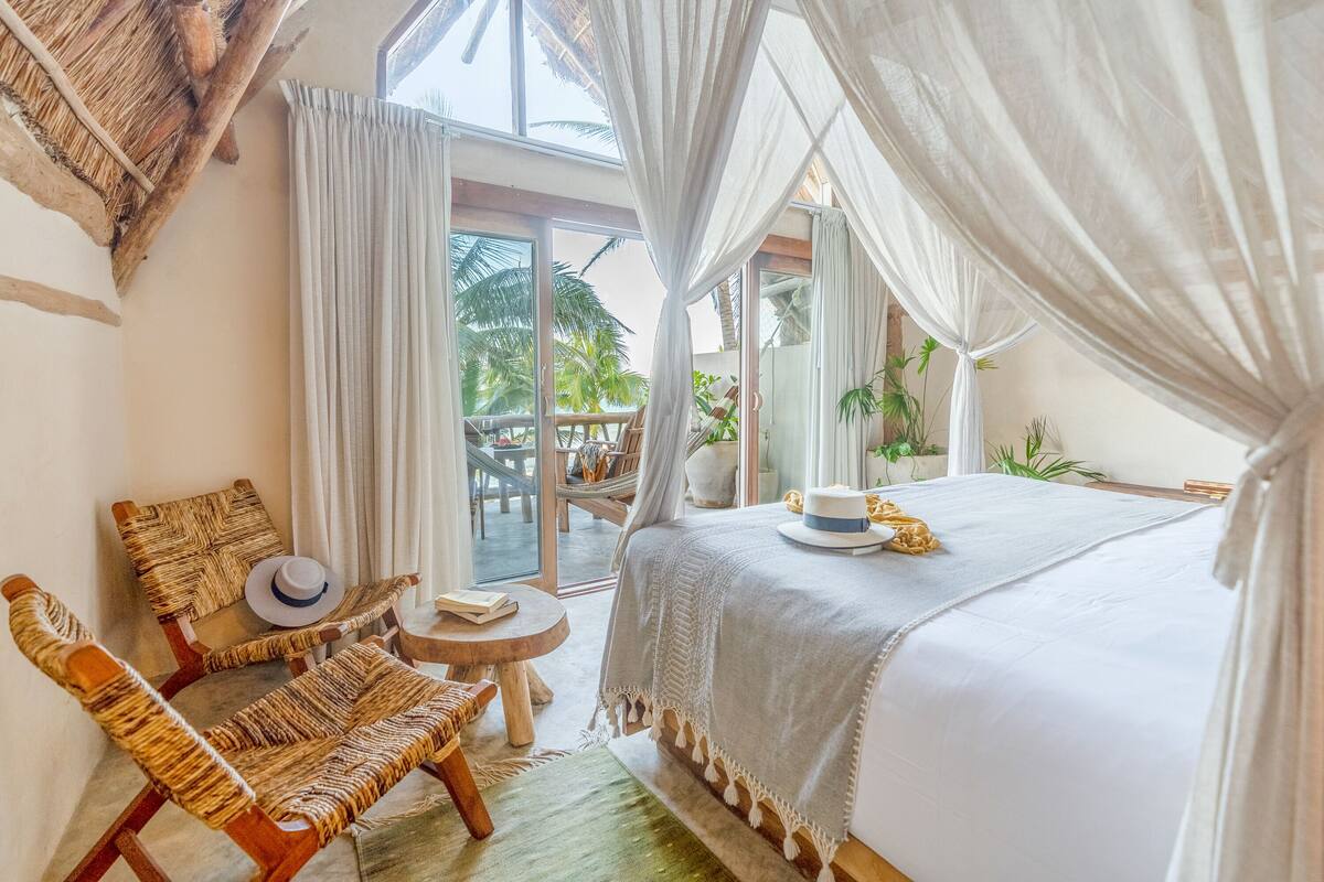 A bedroom with a bed, a chair, a table, and a view of palm trees.
