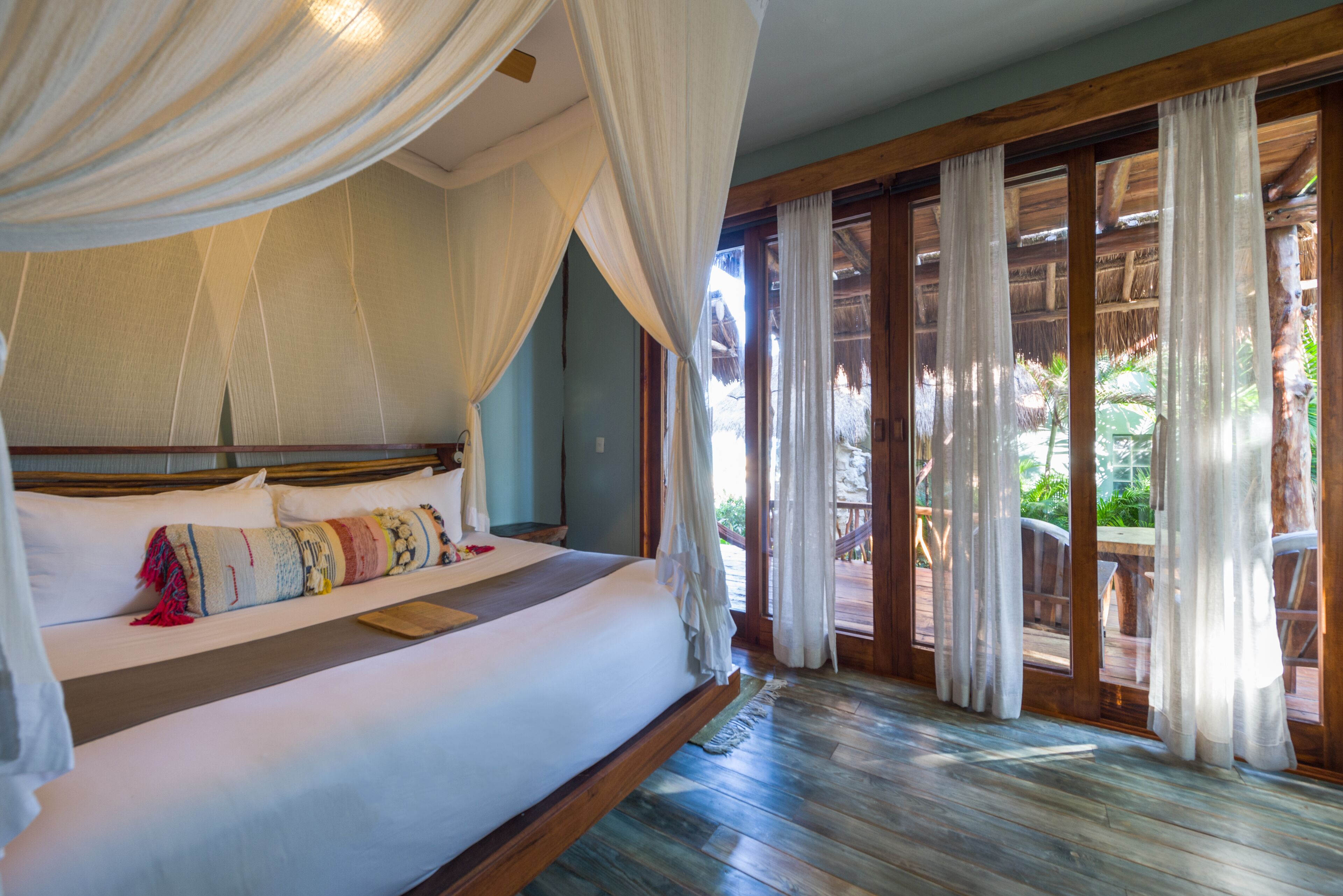A large bed with a canopy, wooden flooring, and a view of the outdoors through a glass door.