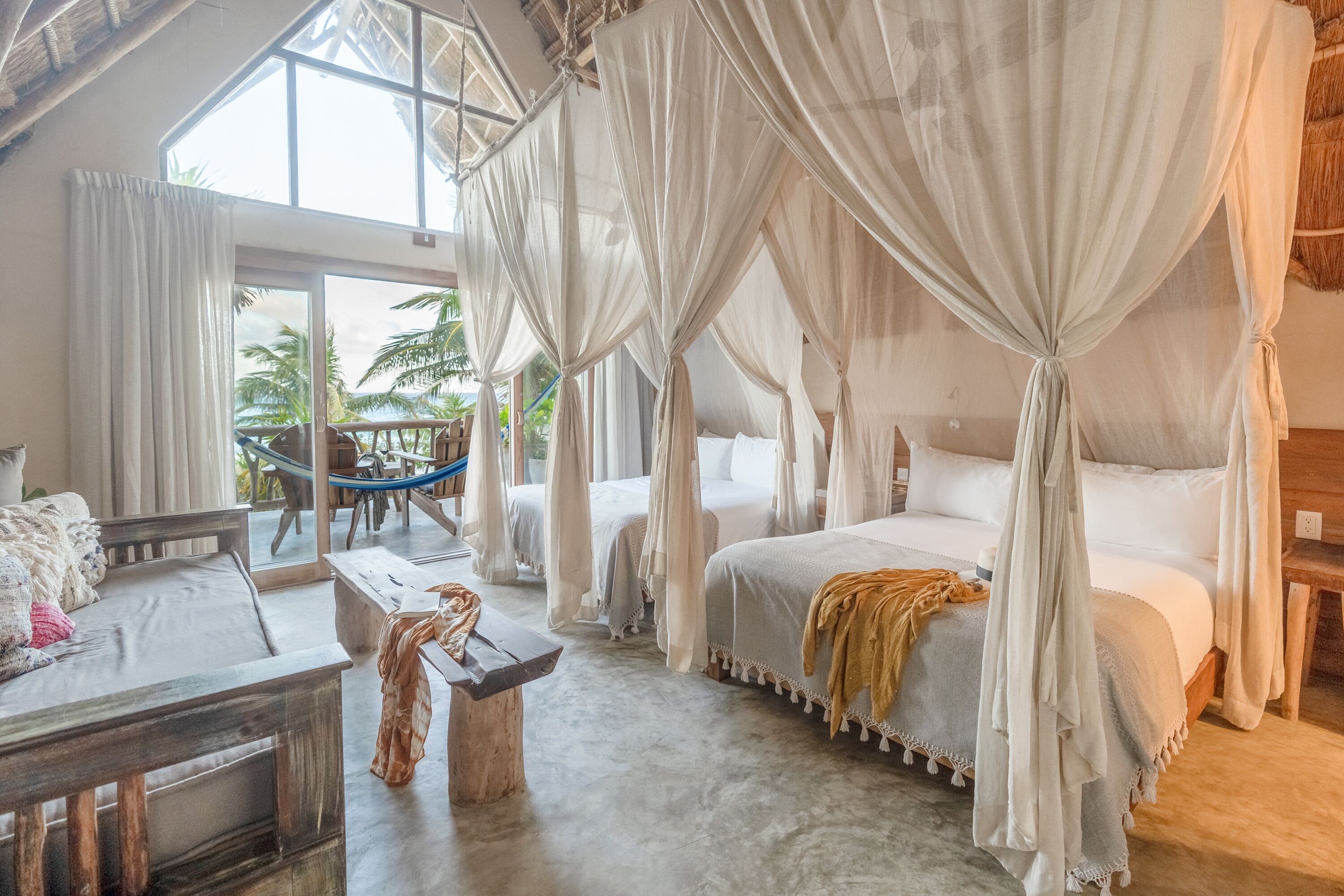 A room with a large bed, a smaller sofa, a wooden bench, and a dining table.