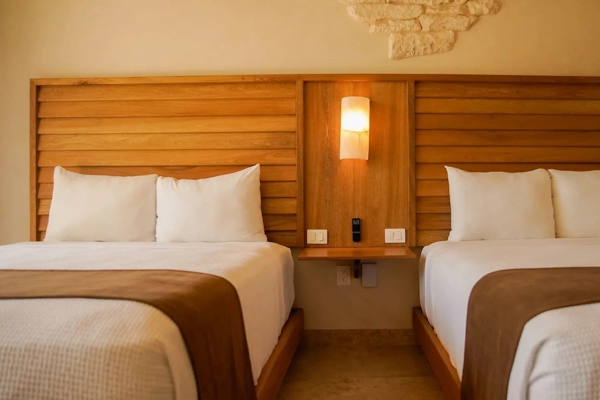Quarto de hotel com duas camas, cabeceiras de madeira, luminária fixada na parede e uma pequena estante.