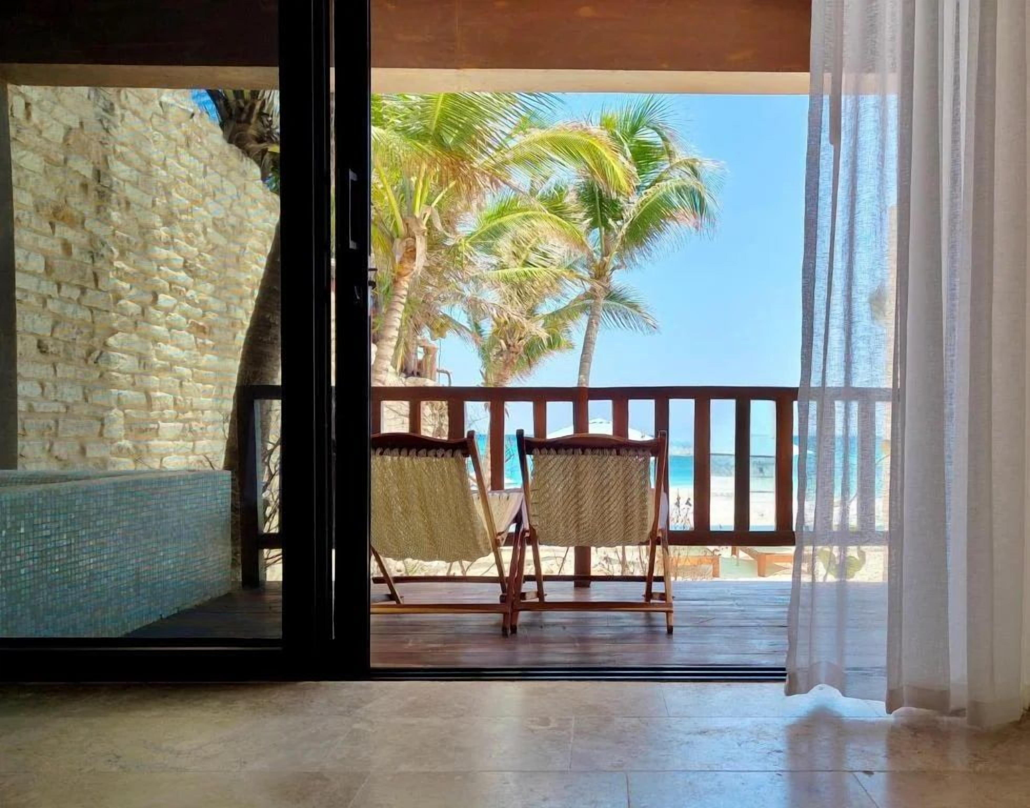 A room with a view of the beach, a stone wall, and two wicker chairs.