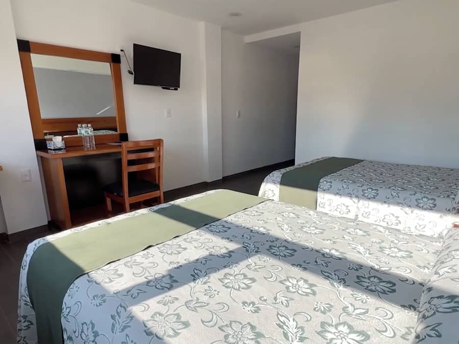 A hotel room with a desk, a mirror, a television, two beds with floral bedding, and a small table with fruits and a glass.