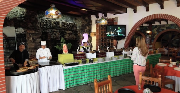 Um restaurante com parede de pedra, mesa de buffet e tela de televisão exibindo uma banda.