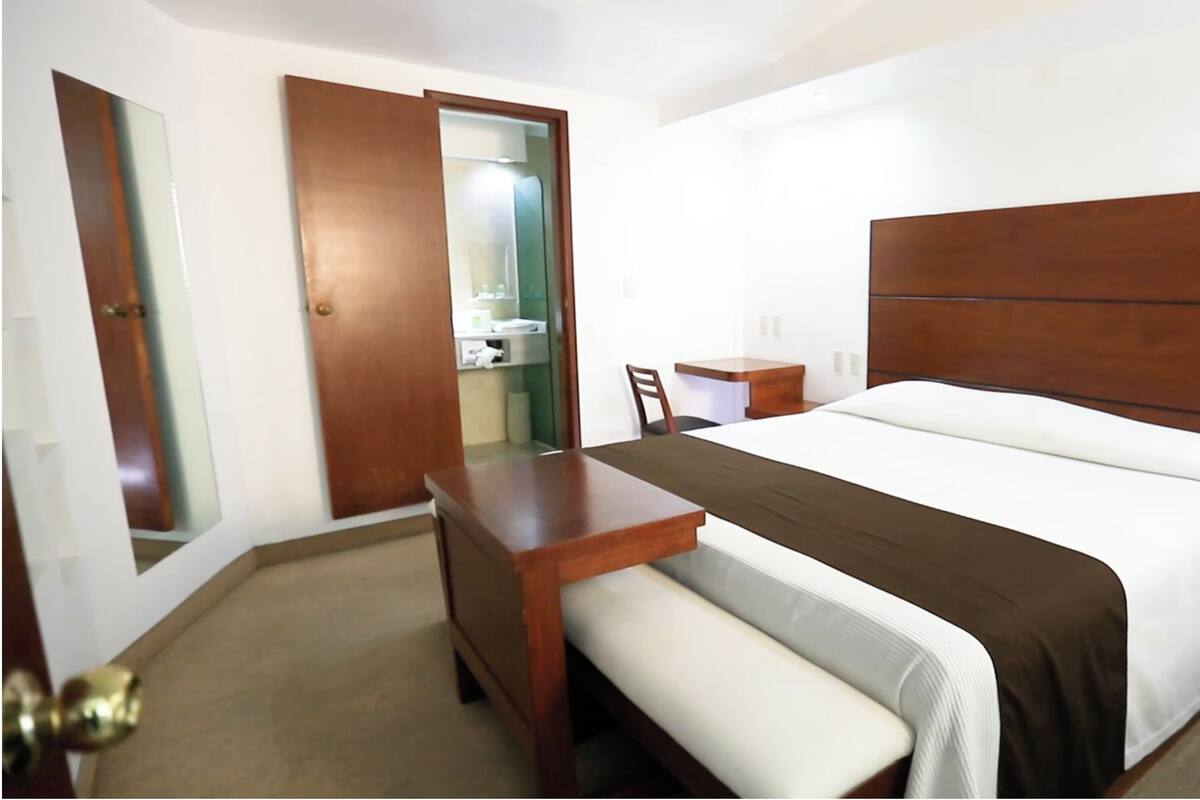 A hotel room with a bed, bedside table, chair, and a small desk with a telephone.