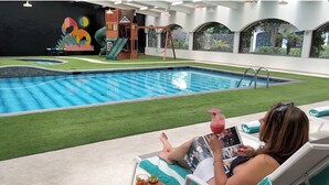 Un espace piscine avec un toboggan et une structure de jeux, une personne lisant un magazine et une fresque murale.