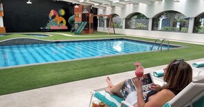 A pool area with a slide and a play structure, a person reading a magazine, and a mural on the wall. - Hotel GS Jerocs Tlaxcala (Tlaxcala)