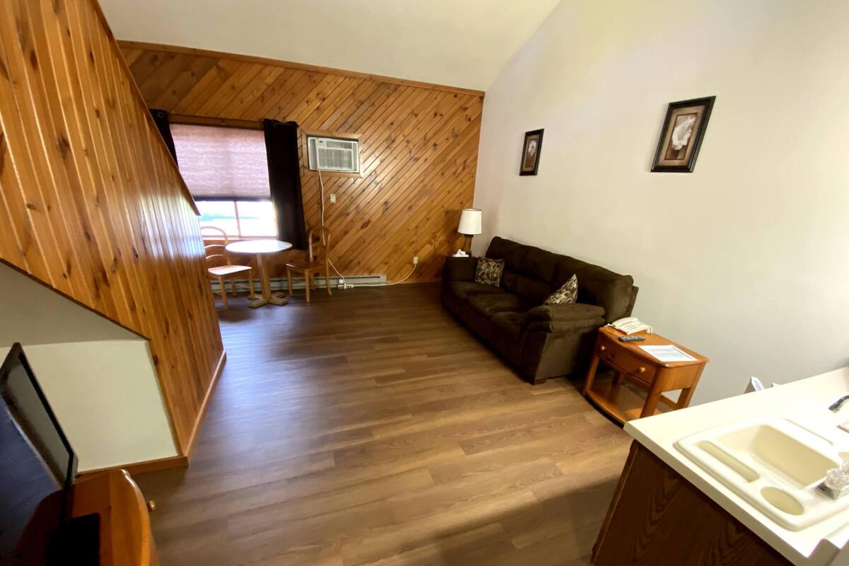 A cozy room with a wooden floor, a sofa, a small table with a lamp, a chair, and a window with curtains.