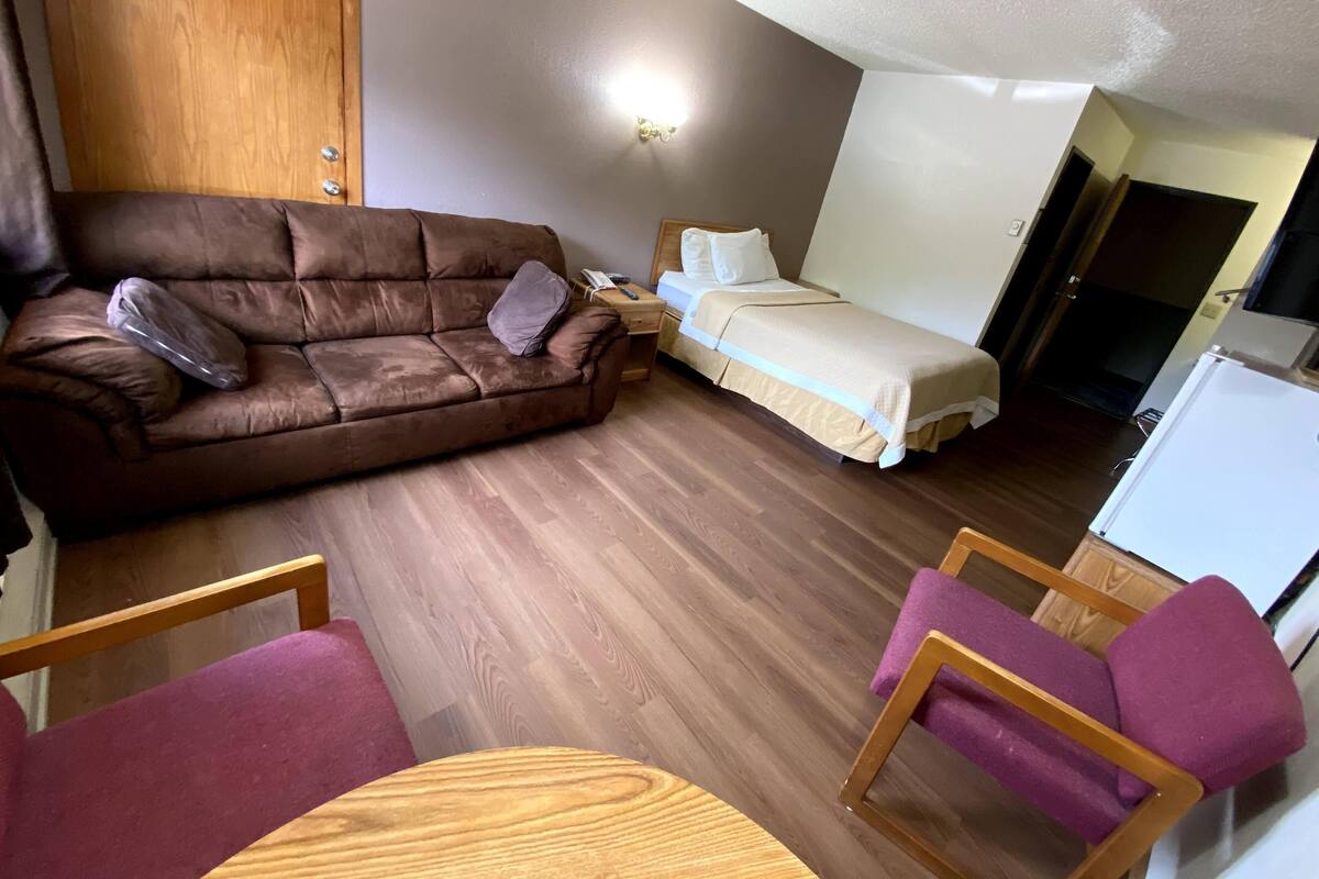 A compact room with a brown sofa, a bed, a small wooden table, and a purple chair.